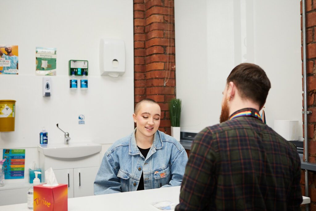 A fem-presenting person in denim jacket talking to LGBT Foundation sexual health officer calmly. Focus on the service user.