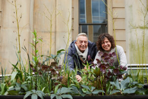 Two older gay and lesbian people in a garden, smiling behind plants.