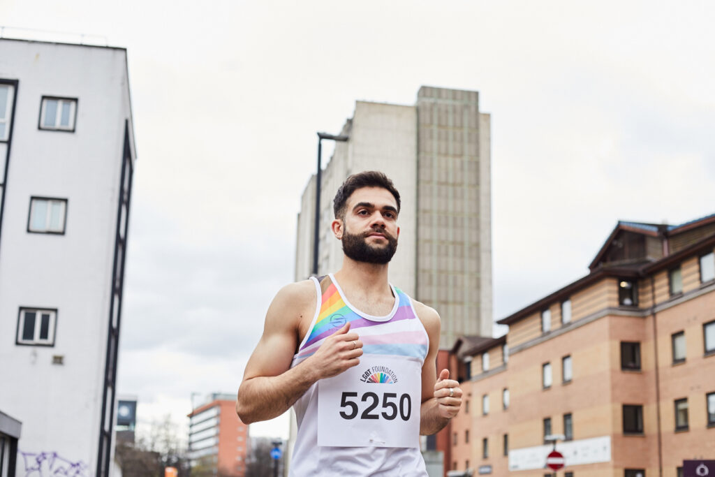 A young bearded bisexual man is shown running down a street in Manchester wearing LGBT foundation's fundraisers running vest