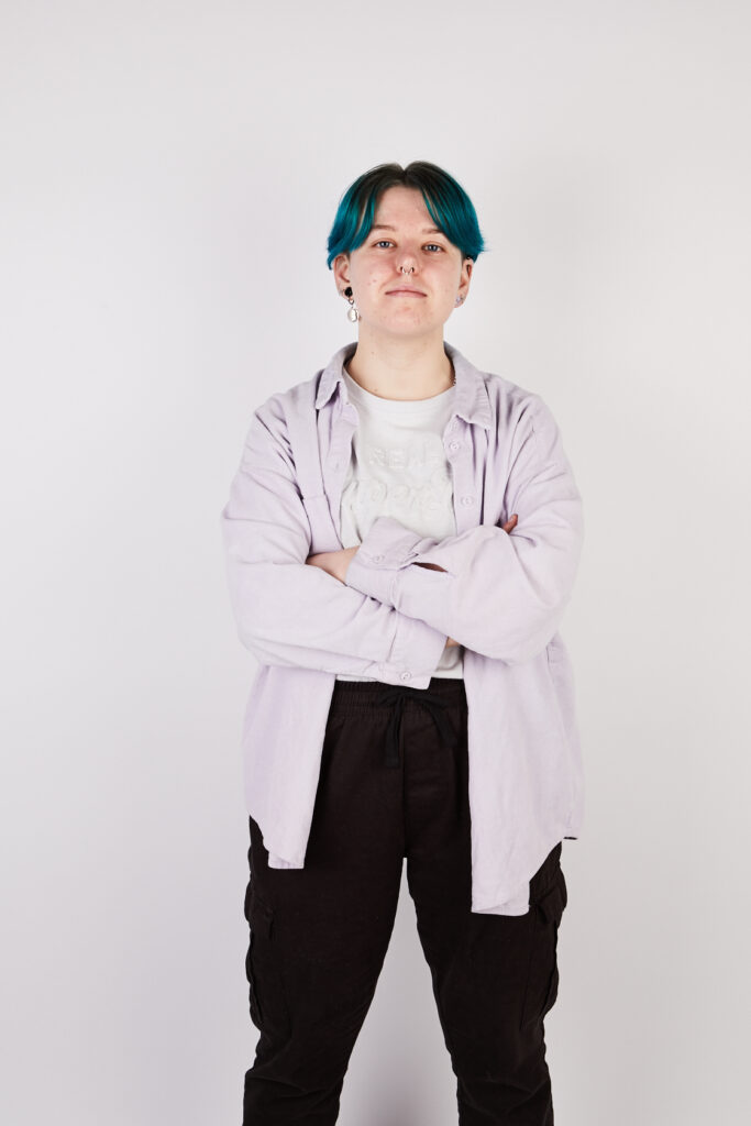 Non-binary person with short blue hair standing confidently with their arms crossed. Portrait.