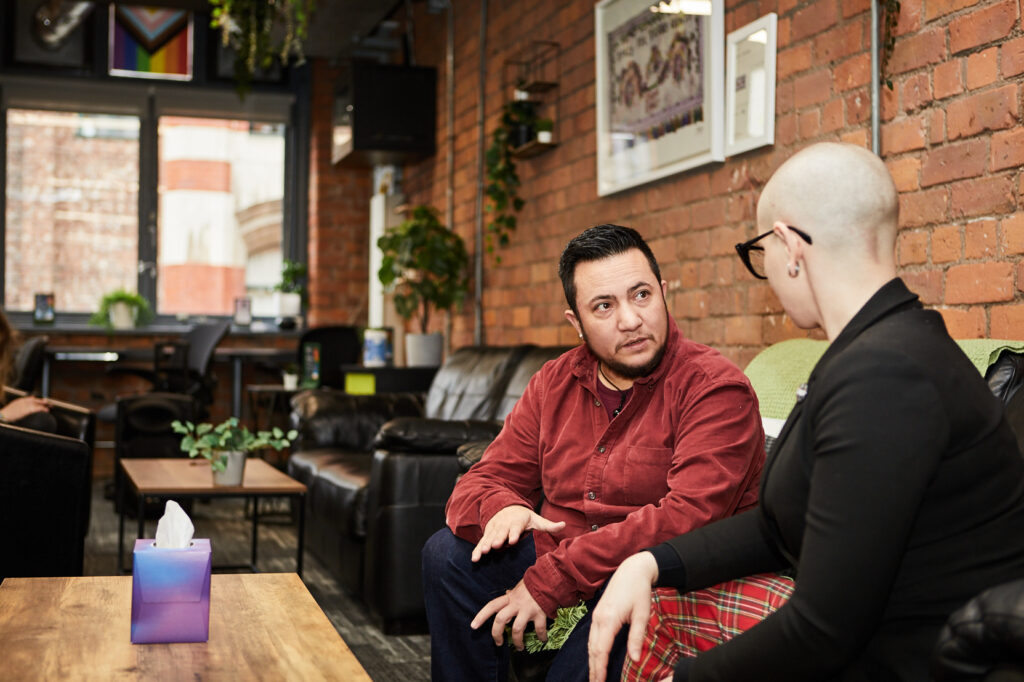 A masc-presenting person talking to a fem-presenting person on sofa, looking concerned.