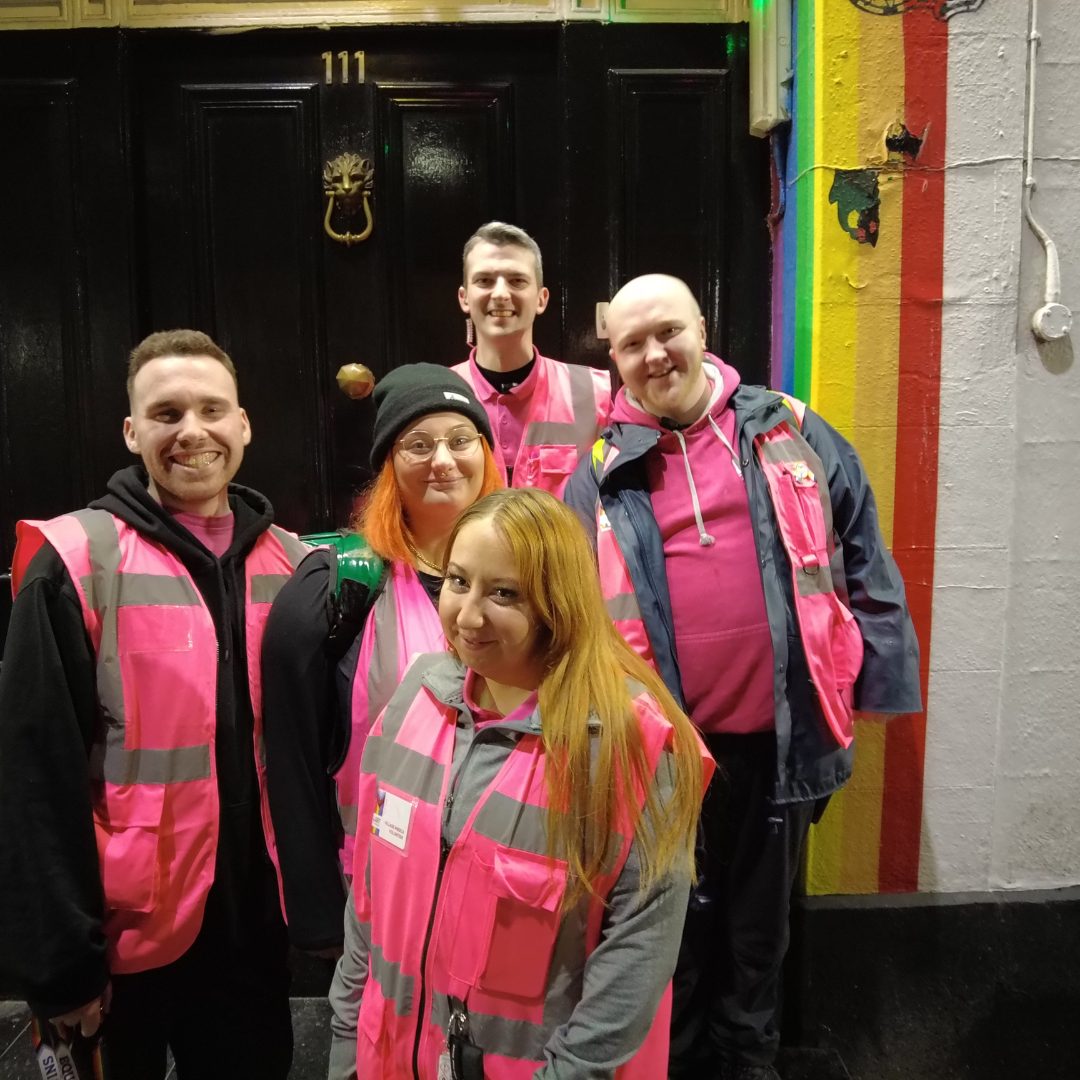 Five village angels in pink hi vis vests standing in front of wooden door and rainbow arch, smiling at camera. Group portrait.