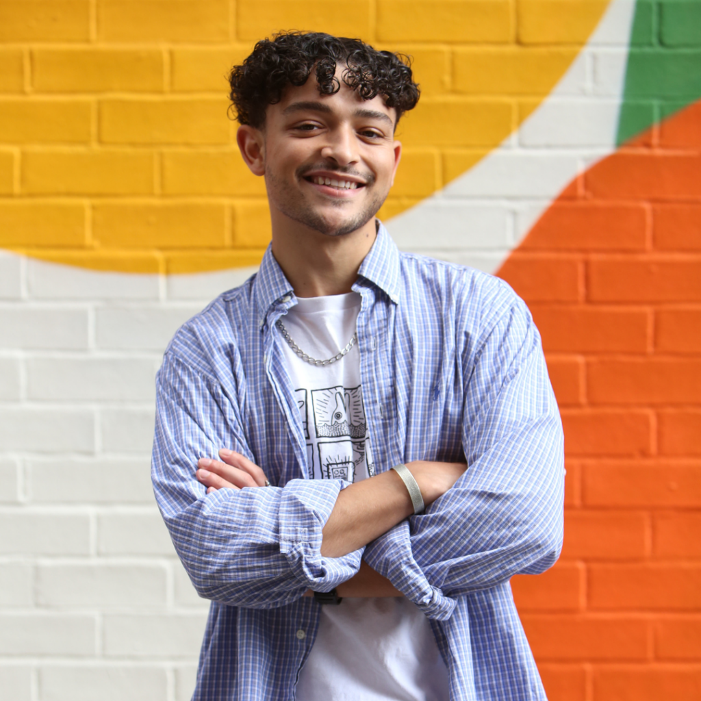 Young brown man with curly hair in light blue shirt standing in front of painted wall, arm crossed in front of chest, smiling at camera. Portrait.