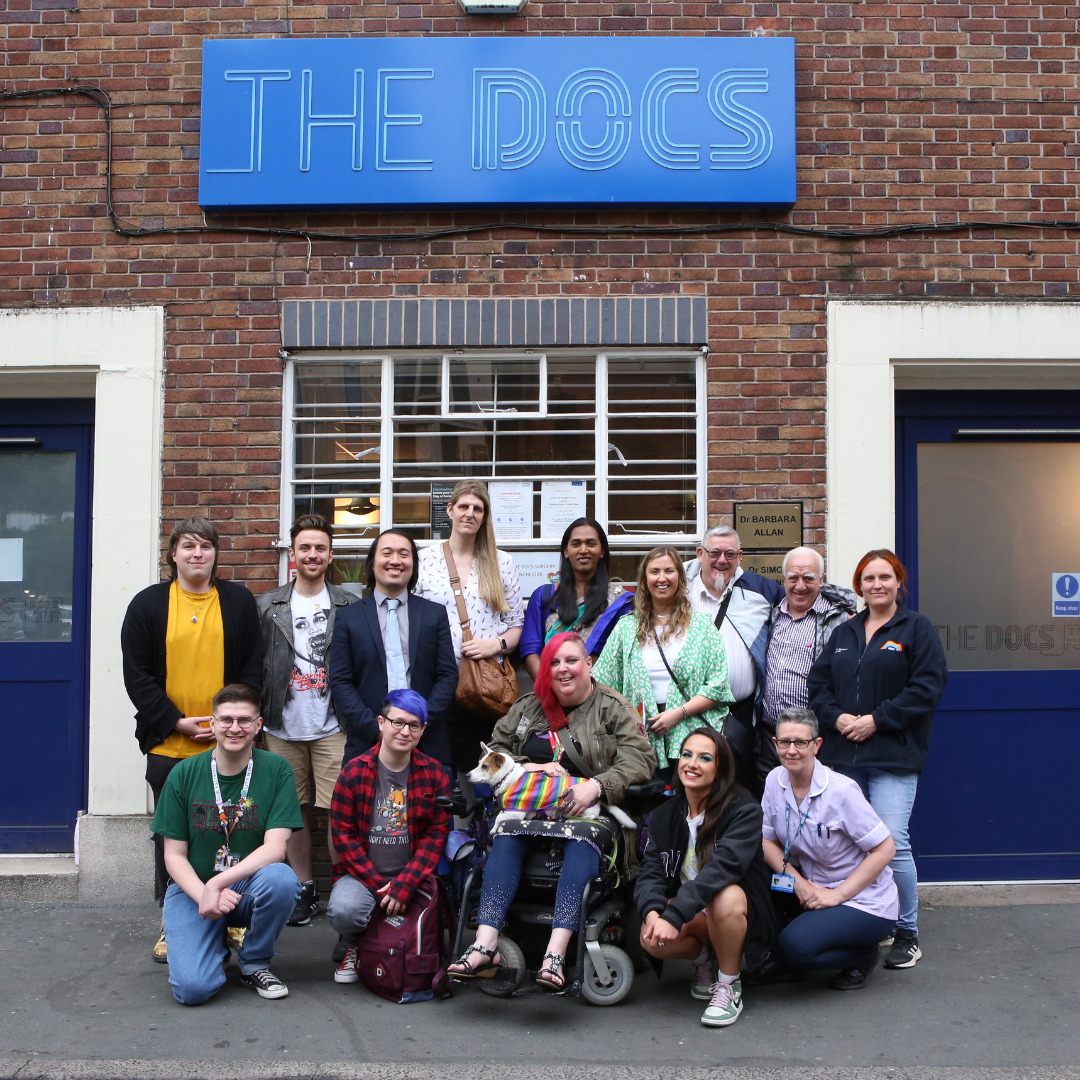 A large group of people in front of a brick wall. Large blue plate hanging on wall reading 'the Docs'. Group portrait of NHS doctors, LGBT Foundation employees and volunteers, and a puppy in rainbow clothes sitting on lap of the person with red hair in the middle.