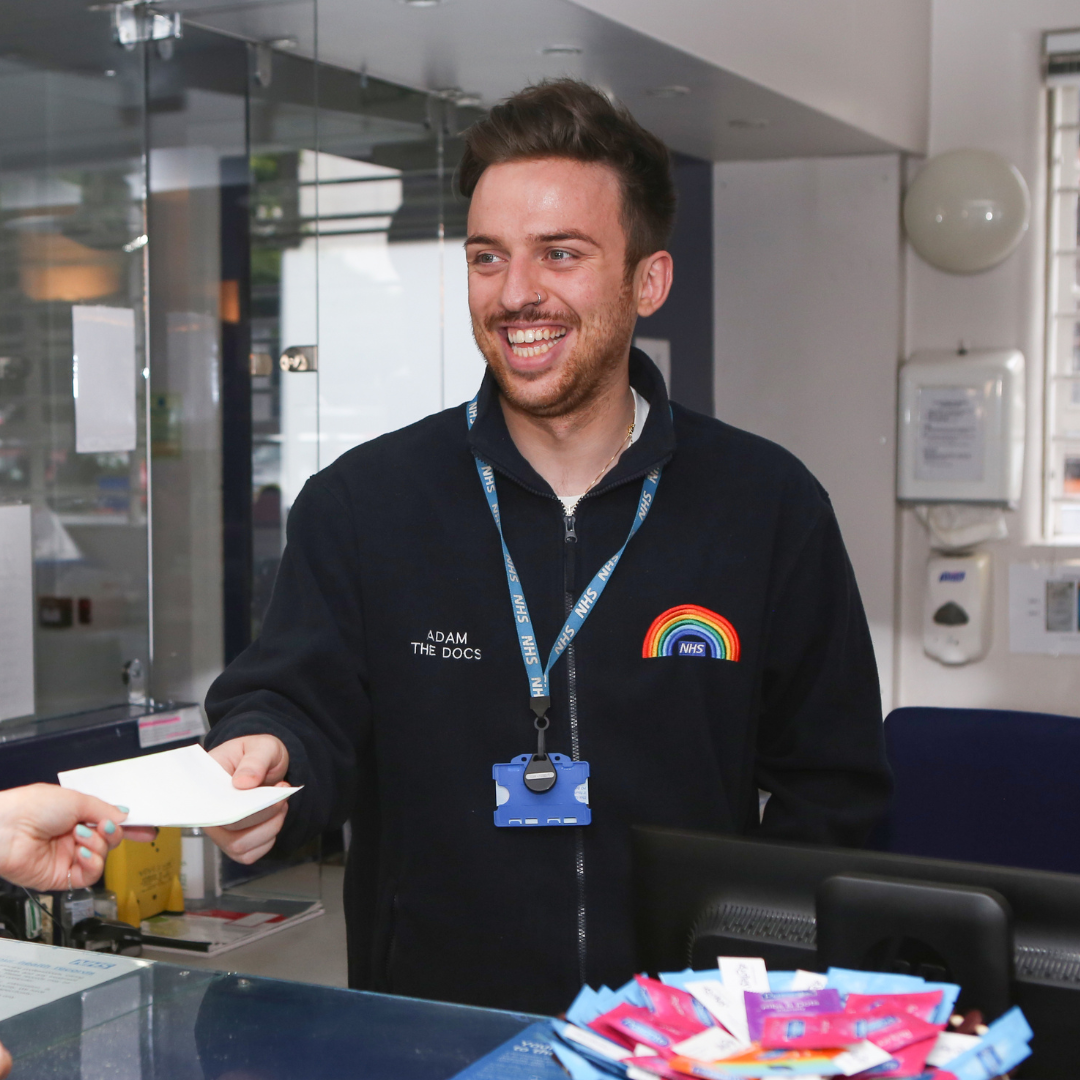 An NHS employee handing a piece of paper to a client with a big smile, wearing a uniform with a rainbow on it.