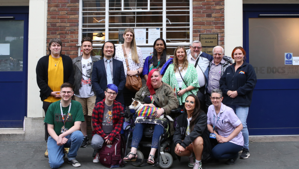 A large group of people in front of a brick wall. Group portrait of NHS doctors, LGBT Foundation employees and volunteers, and a puppy in rainbow clothes sitting on lap of the person with red hair in the middle.