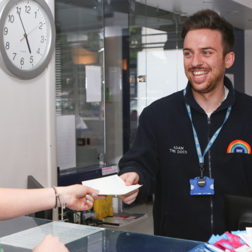 An NHS employee handing a piece of paper to a client with a big smile, wearing a uniform with a rainbow on it.