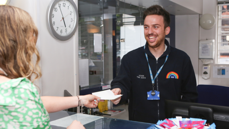 An NHS employee handing a piece of paper to a client with a big smile, wearing a uniform with a rainbow on it.