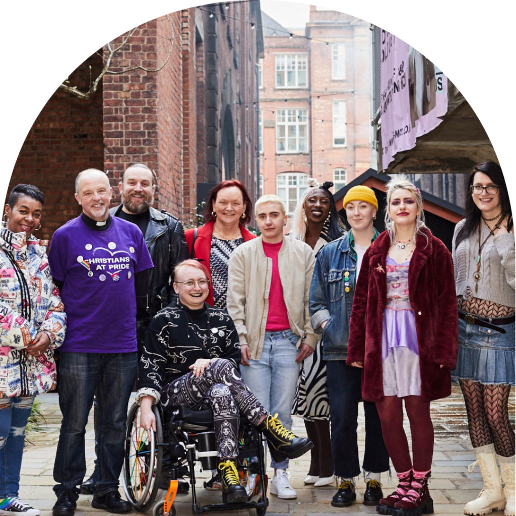 A group of diverse people standing in an alleyway, smiling at camera. In the middle, a fem-presenting person sitting on wheelchair. On the side, people of different gender identities standing in a line. Group portrait.