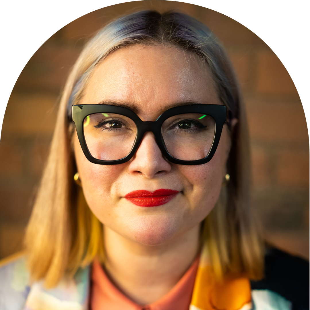 Non-binary femme presenting person with blonde hair wearing thick black framed glasses smiling at camera. Portrait.