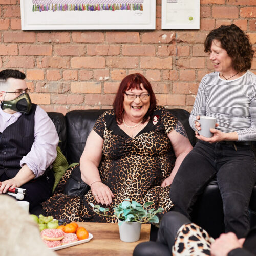 Two fem-presenting and one masc-presenting people sitting on sofa with a table in the front. All smiling and listening to a person outside the frame talking. Table full of fruit, snacks and coffee cups.