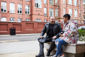 White man in leather outfit and black woman in colourful winter coat talking and smiling. Both sitting on a bench on the side of main road.