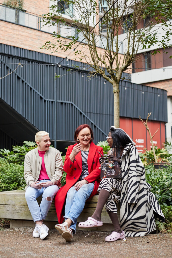 Three People sitting on bench under tree. Two fem-presenting people on the left and one Mac-presenting on the left. All talking and laughing.