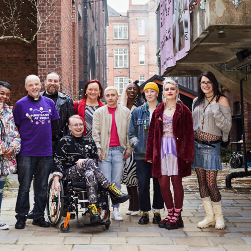 A group of diverse people standing in an alleyway, smiling at camera. In the middle, a fem-presenting person sitting on wheelchair. On the side, people of different gender identities standing in a line. Group portrait.