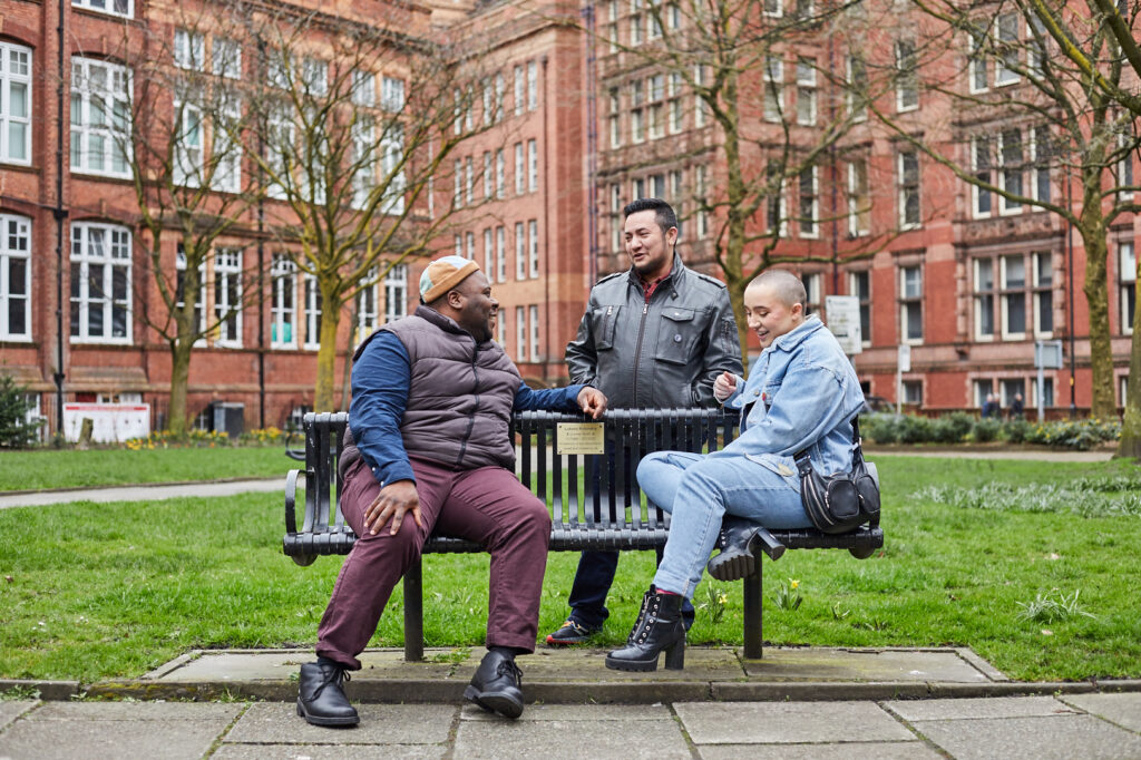 Three friends chatting and smiling around a bench in Sackville gardens.
