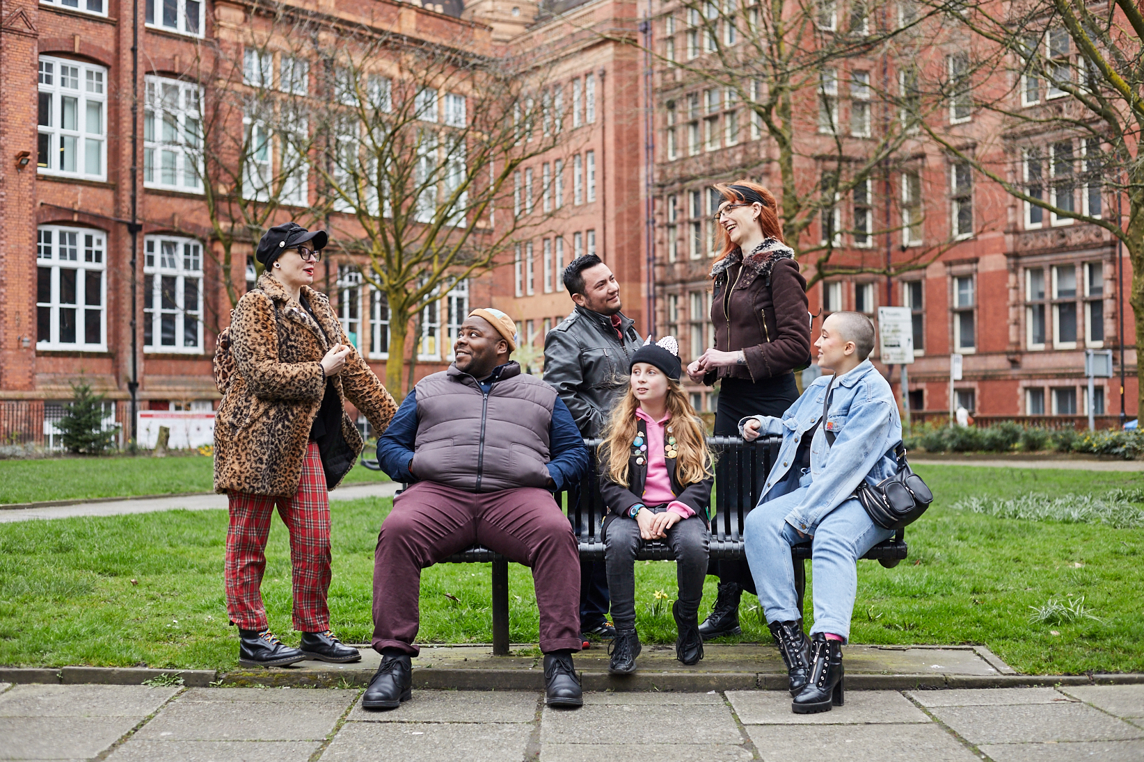 Five people talking around a bench in garden. A black man, a young child in beanie with cat ears and a fem-presenting person in denim jacket sitting, while a fem-presenting person in fur-coat, a trans man and a trans woman standing behind, all talking.