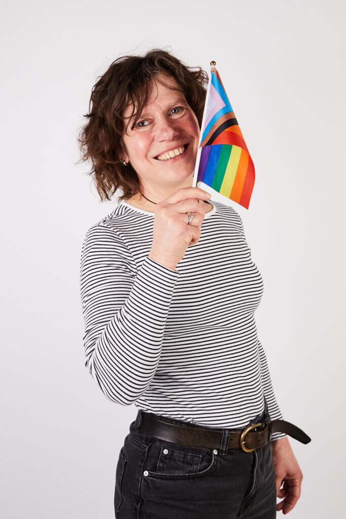 Older woman in striped t-shirt and jeans holding a pride flag, smiling at camera. Portrait.