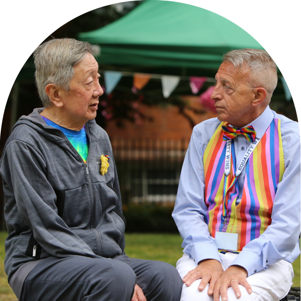 Two gay older men, one east asian in grey hooddie and the other white in rainbow waistcoat, facing each other, talking.