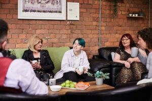 Two trans women, a non-binary person and an older woman sitting around a table, chatting and drinking tea. A tray full of fruits and donuts on table.
