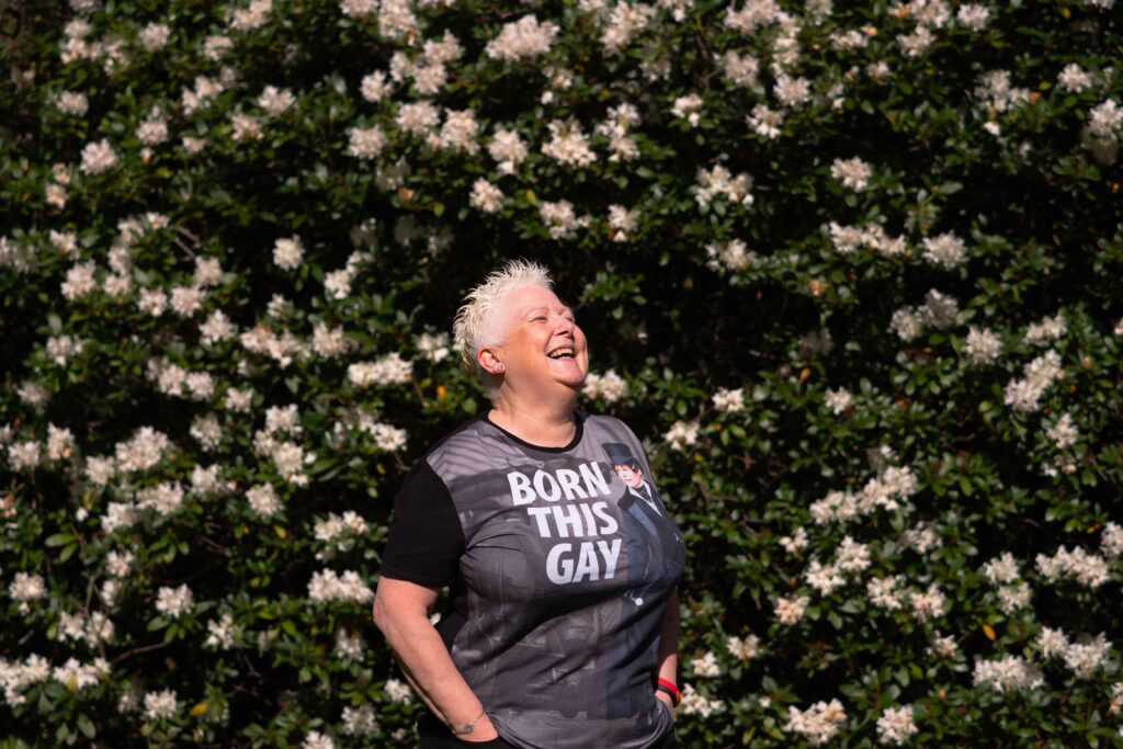 Older fem-presenting person wearing t-shirt reading 'Born this Gay' in short blond hair smiling happily in the sun, facing sideway. Background of beautiful bushes with lots of white flowers in them. Portrait.