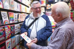 Two older gay men with white hair selecting cards in card shop for Pride. Both looking at each other and smiling.