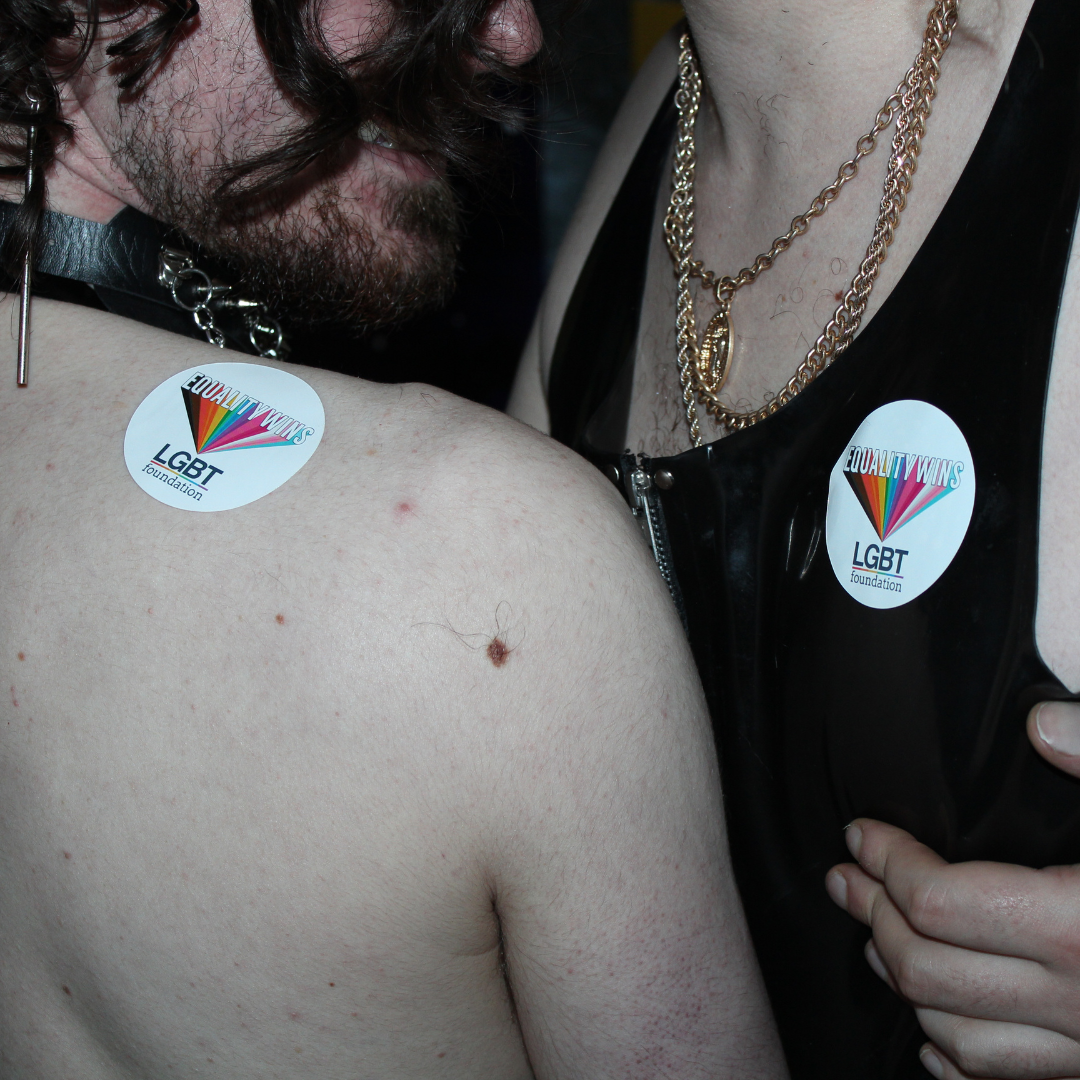Two men holding each other. One with metal chain on neck and the other in leather top. Both have stickers reading 'LGBT Foundation'. Close-up on chests.