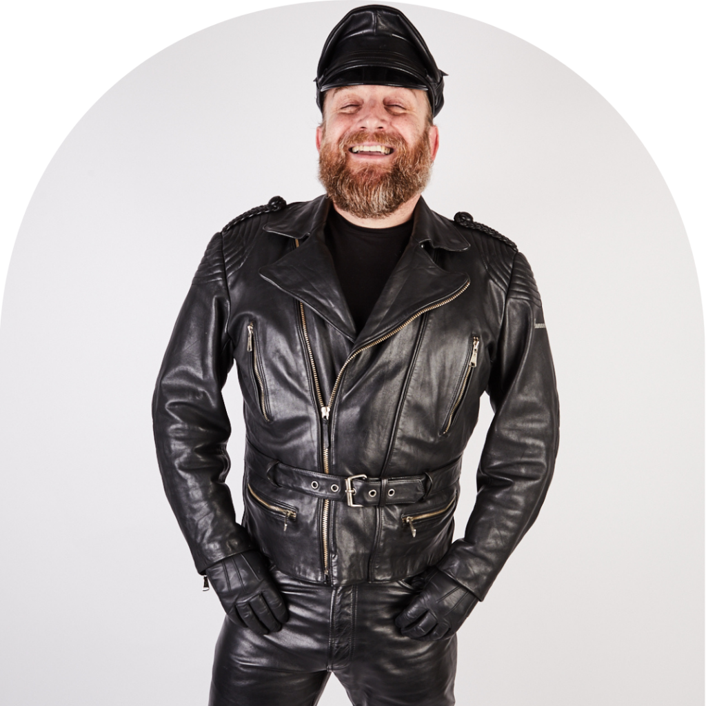 Man in leather jacket, trousers, gloves and hat facing camera, smiling.