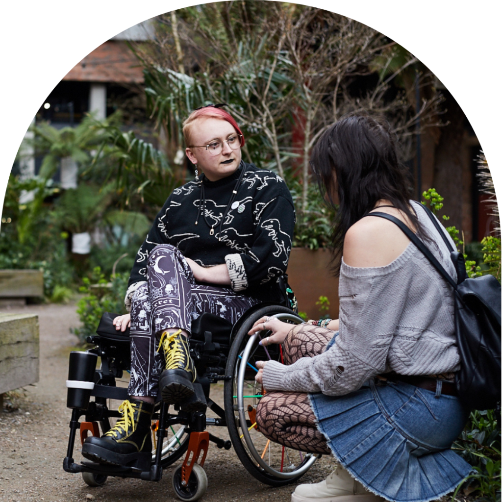 A fem-presenting person on wheelchair talking to a trans woman, looking concerned.