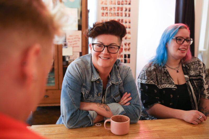 Woman in denim jacket with arms crossed smiling and talking to man with back to camera. Empties coffee mug on table. Woman in pink and blue hair sitting next to them.