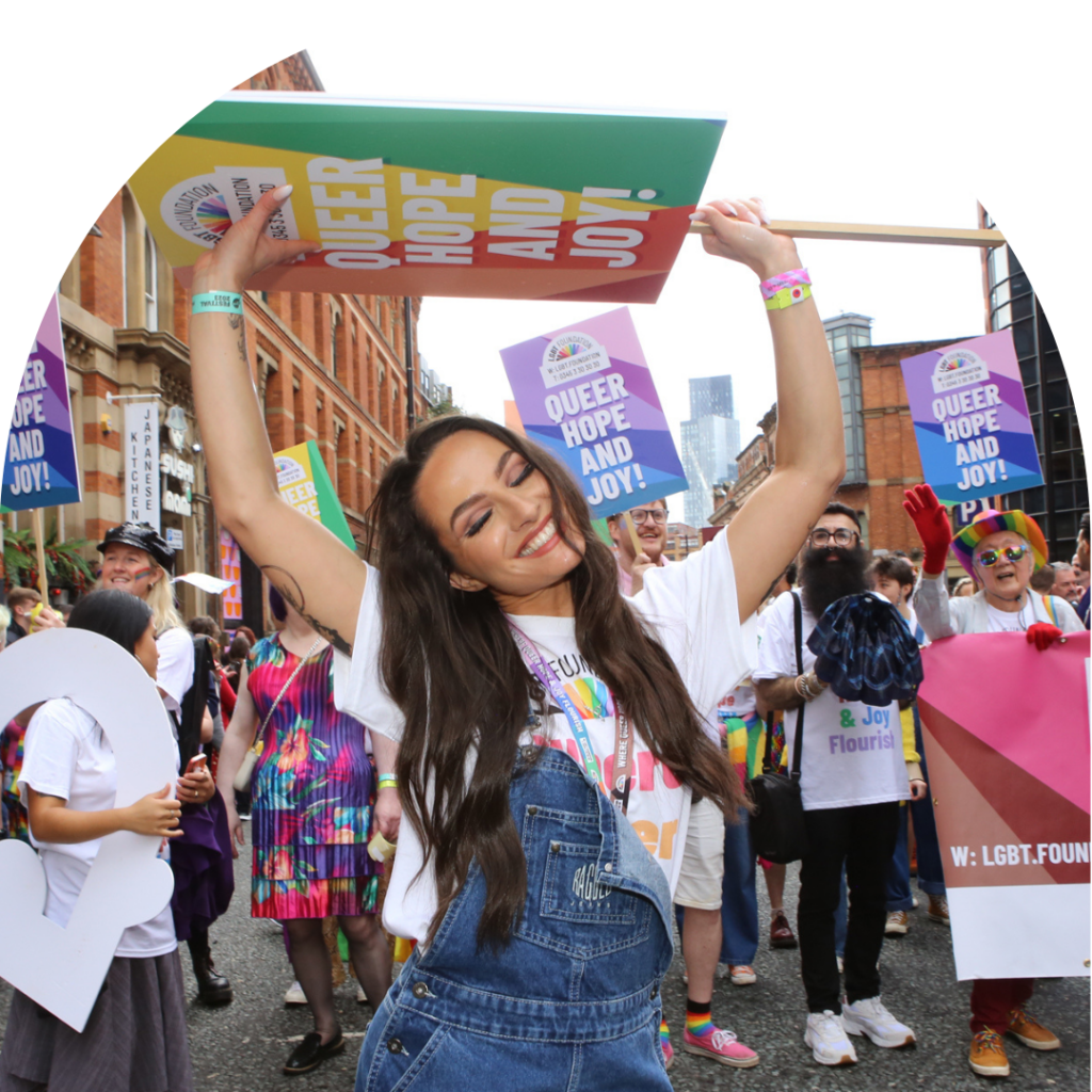 A young femme presenting bi woman stands holding a placard while smiling at the front of a pride parade this image is in an arch frame