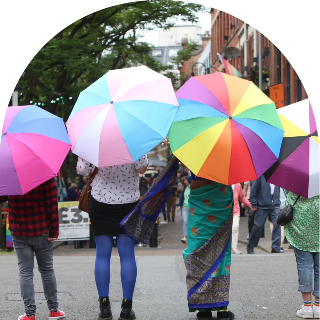 Four people stand with their backs facing the camera, they each are wearing different and unique items of clothing, and stand at different heights. You cannot see their heads as they cary umbrellas in colours of sexuality identity flags