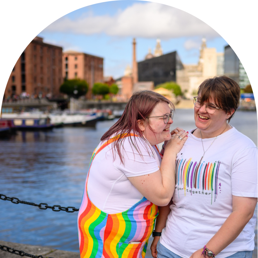 Two queer people, one is femme-presenting woman the other non-binary, in rainbow shirts standing by a river, enjoying the scenic view.