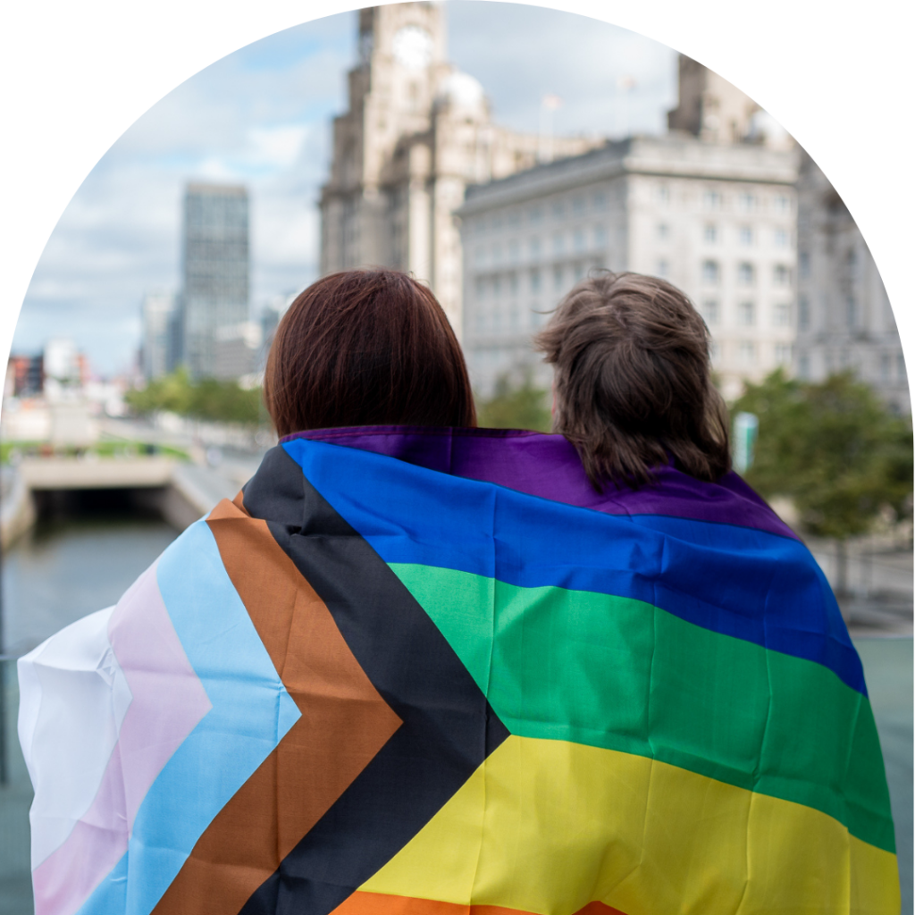 back of two LGBT people stand looking over Liverpool docks with a pride flag over their shoulders 