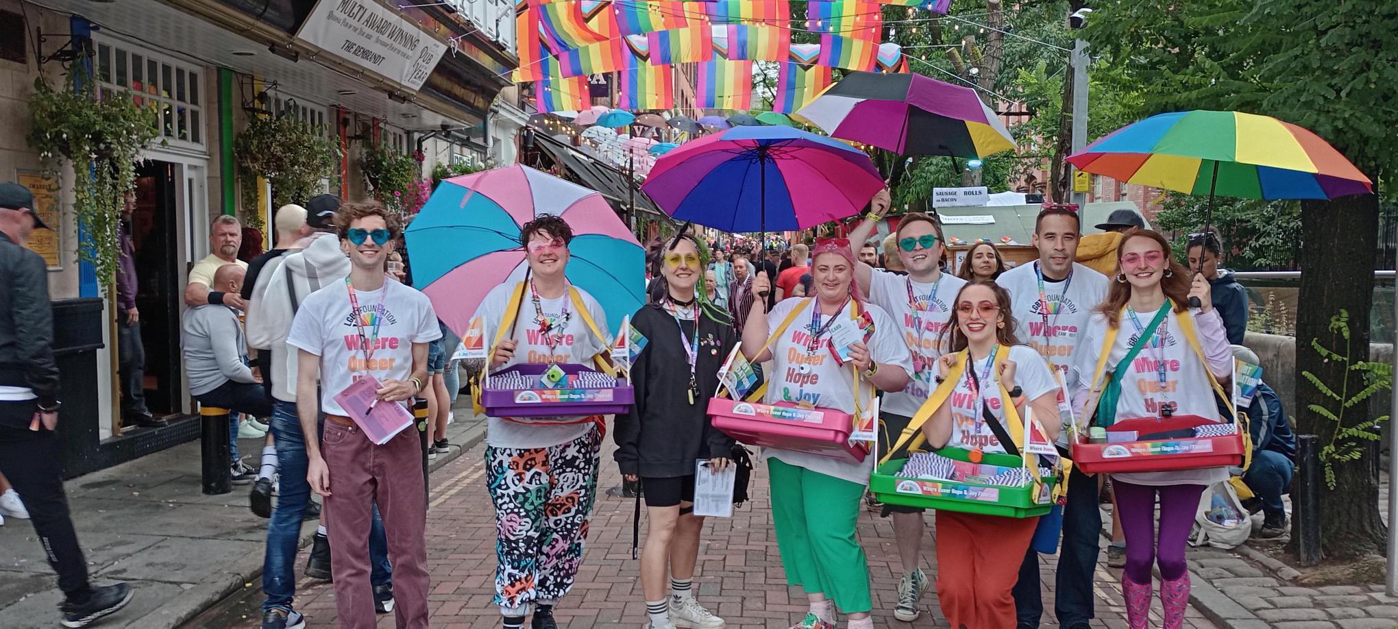 Our sexual health team and their volunteers stand on canal street with their freebies including condoms and lube using their ice cream tray mobile outreach, they're all holding identity based LGBTQ+ flag umbrellas.