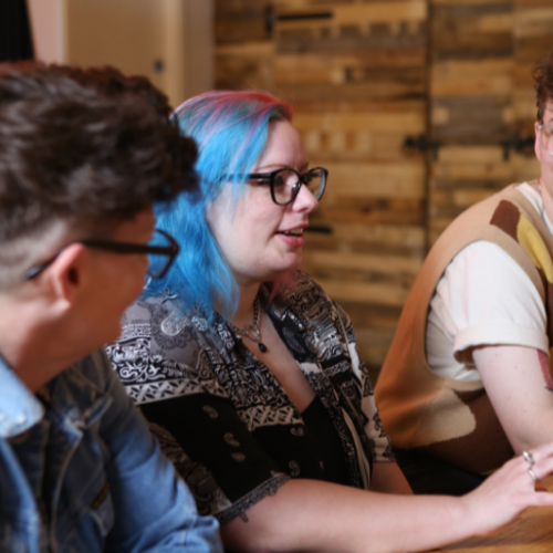 Image shows a group of queer people in discussion around a table