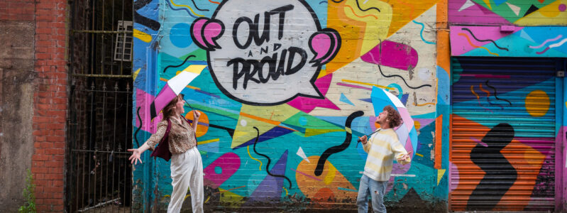 Two LGBTQ+ people celebrating in front of Out and Proud street art