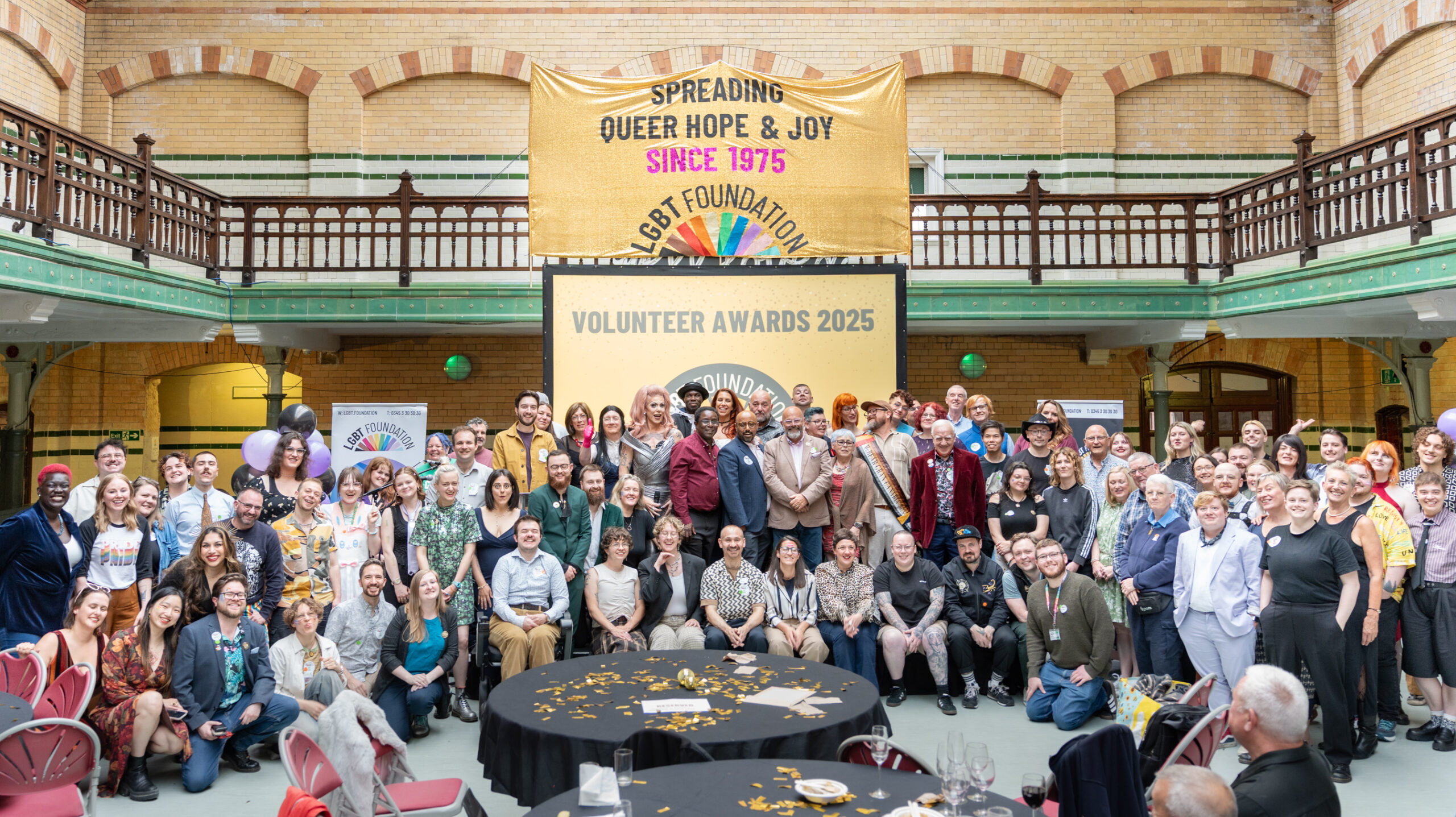 Group photo of staff and volunteers of LGBT Foundation at Victoria Baths, smiling at the camera, with a gold presentation in the background that reads "Volunteer Awards 2025" and a large sequin gold banner above the screen that reads "Spreading Queer Hope & Joy Since 1975" with the LGBT Foundation logo underneath.