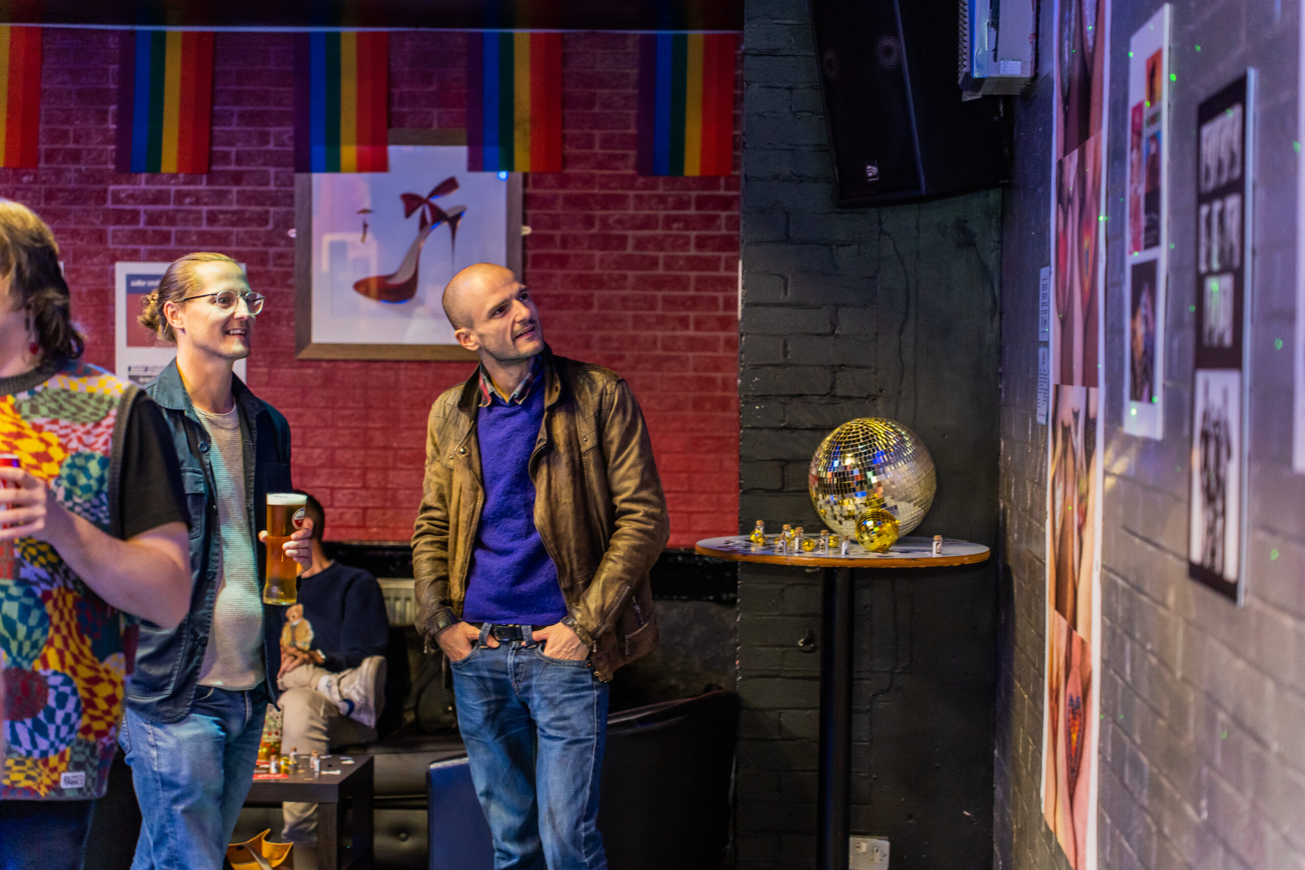 Two masc-presenting people looking at a wall of photographs in the EXXXhibition at Thompsons Arms.