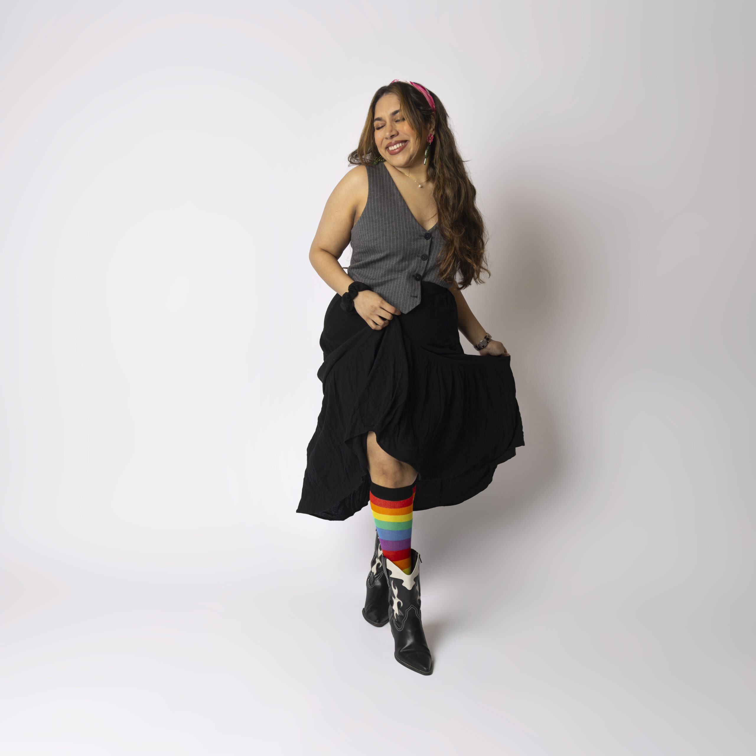 A femme-presenting person smiling and raising their skirt to show off a pair of long rainbow socks.