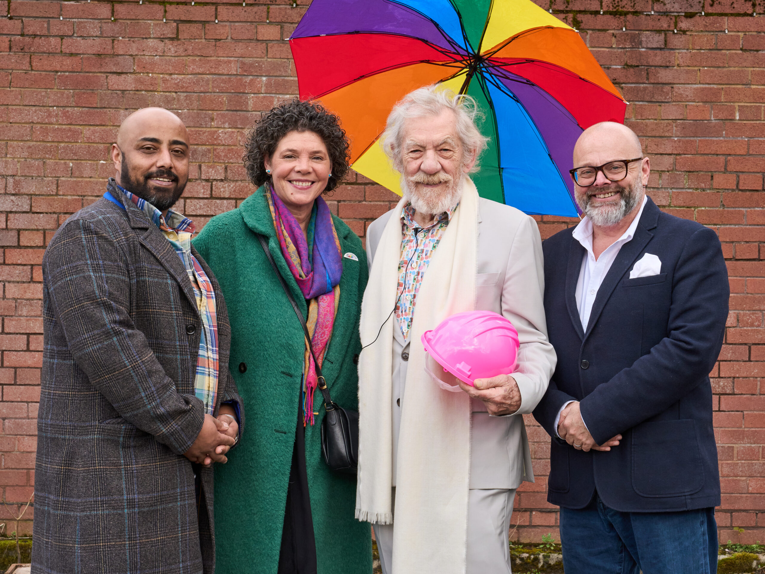 An image of Sir Ian McKellen with LGBT Foundation trustees and CEO, from left to right: Jason Kalugarama, Jax Effiong, Sir Ian McKellen, Paul Martin. The people are smiling in front of a brick wall, with a rainbow umbrella behind Sir Ian McKellen, who is holding a pink hard hat.