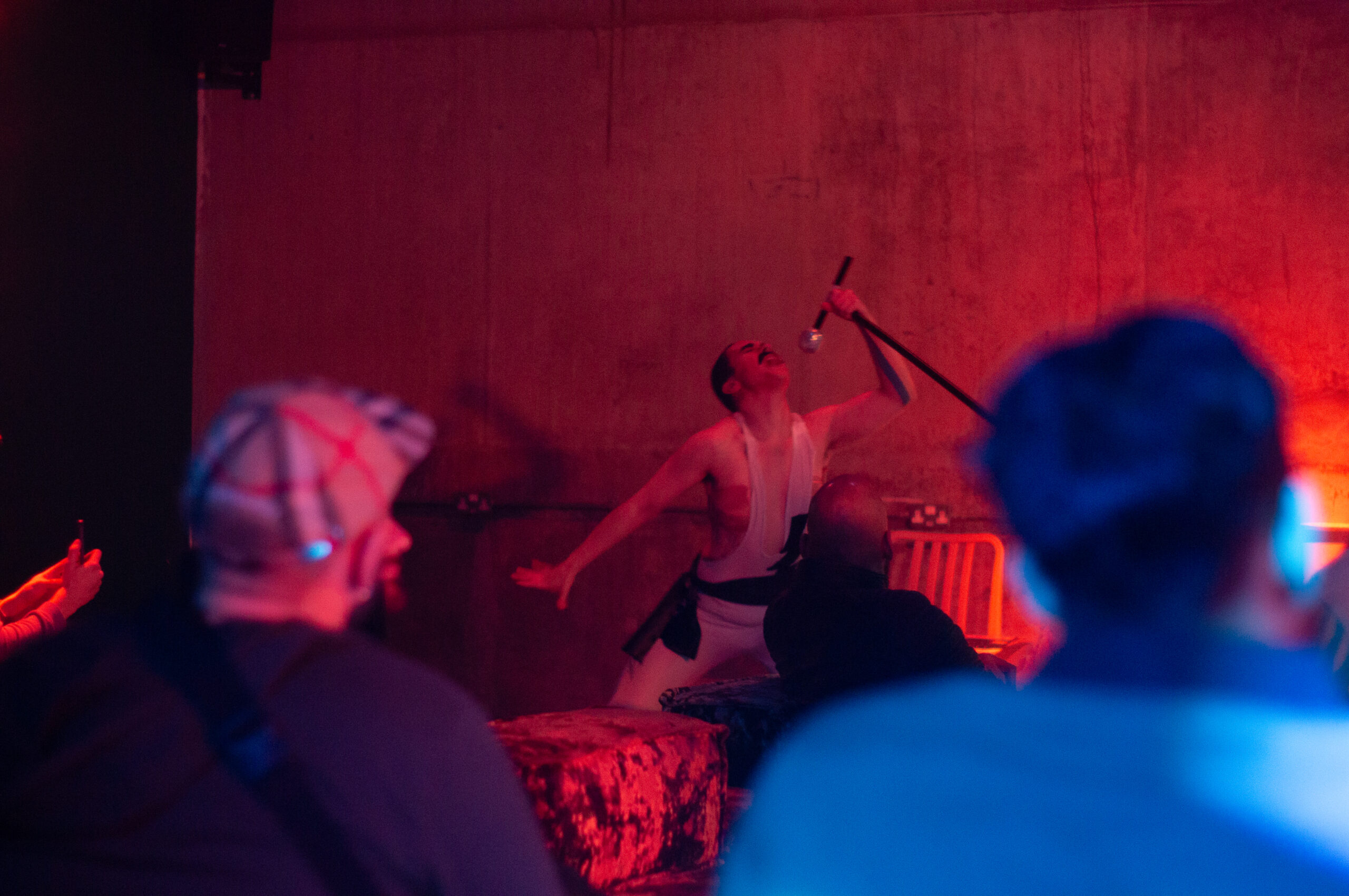 A masc-presenting person singing into a microphone in front of an audience. The lighting in the image is red and dark, indicating a nightlife event.