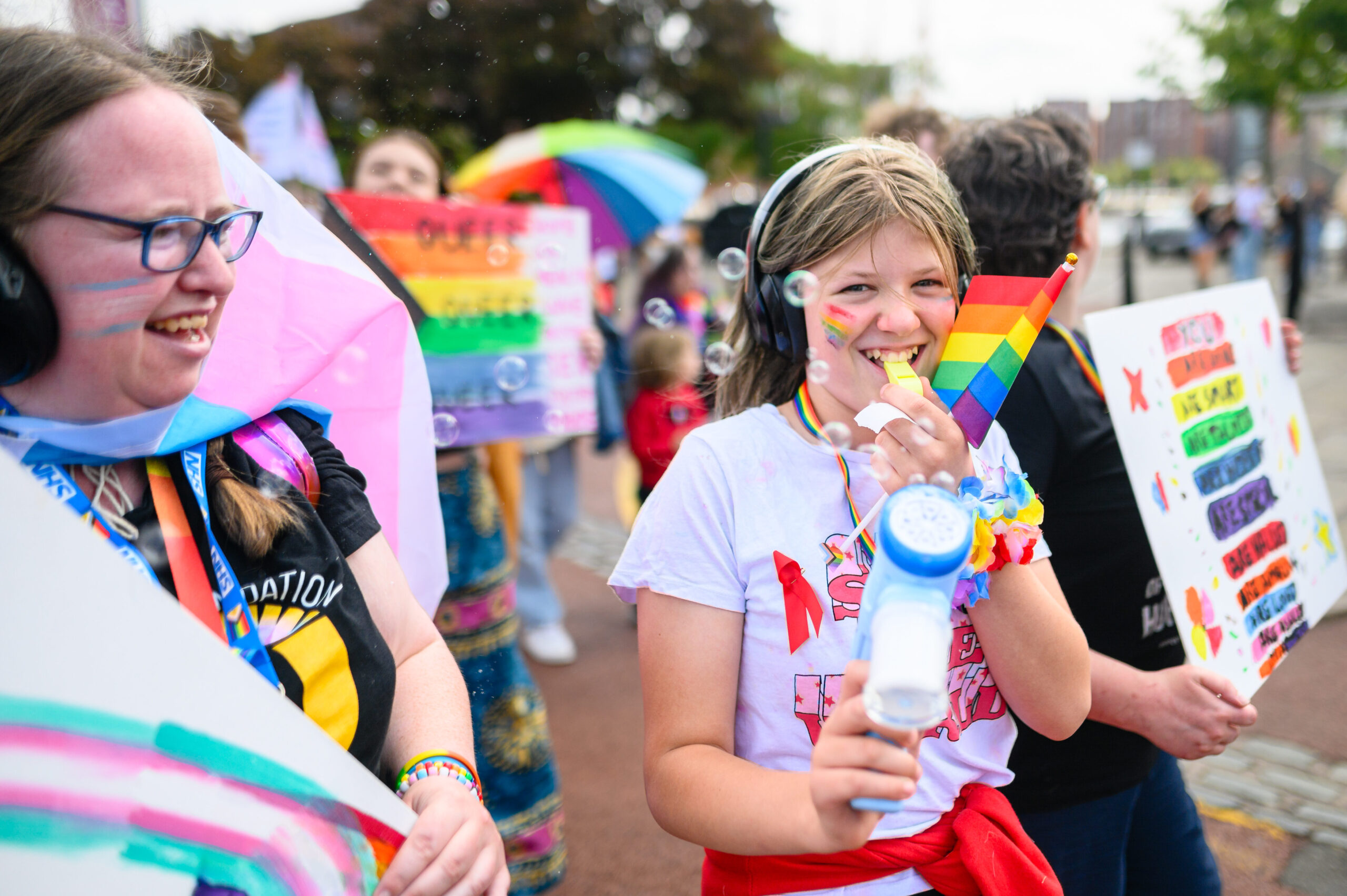 The focus of the image is a young person, smiling to the camera, with headphones on and wearing memorabilia supporting LGBTQ+ causes and with a blue unicorn-themed bubble gun. To their left is another member of the LGBTQ+ community, smiling and wearing glasses and holding a placard with the trans+ colours on.