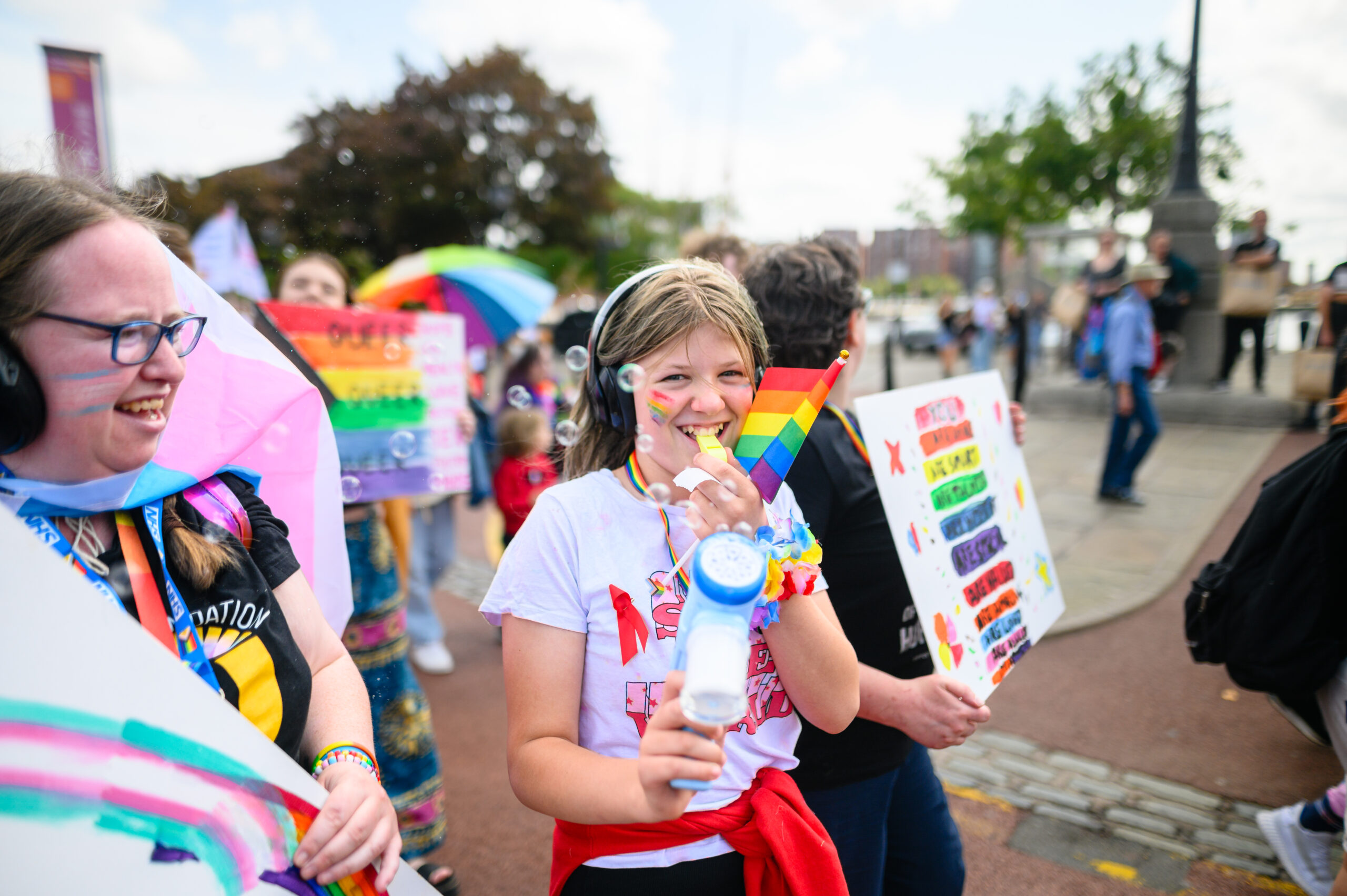 The focus of the image is a young person, smiling to the camera, with headphones on and wearing memorabilia supporting LGBTQ+ causes and with a blue unicorn-themed bubble gun. To their left is another member of the LGBTQ+ community, smiling and wearing glasses and holding a placard with the trans+ colours on.