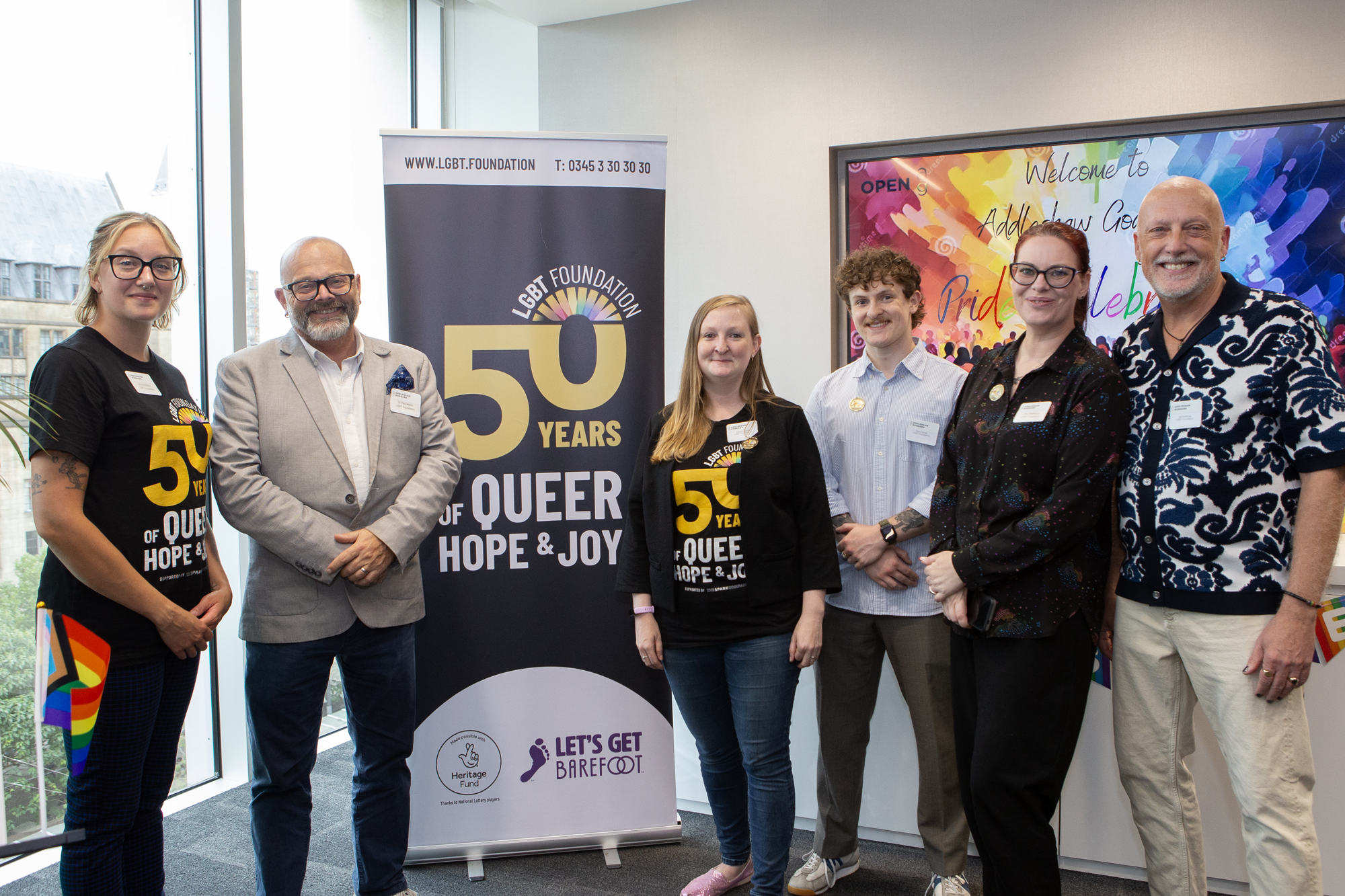 LGBT Foundation staff members smiling to the camera at a corporate event, with a 50 Years of Queer Hope and Joy banner in the centre.