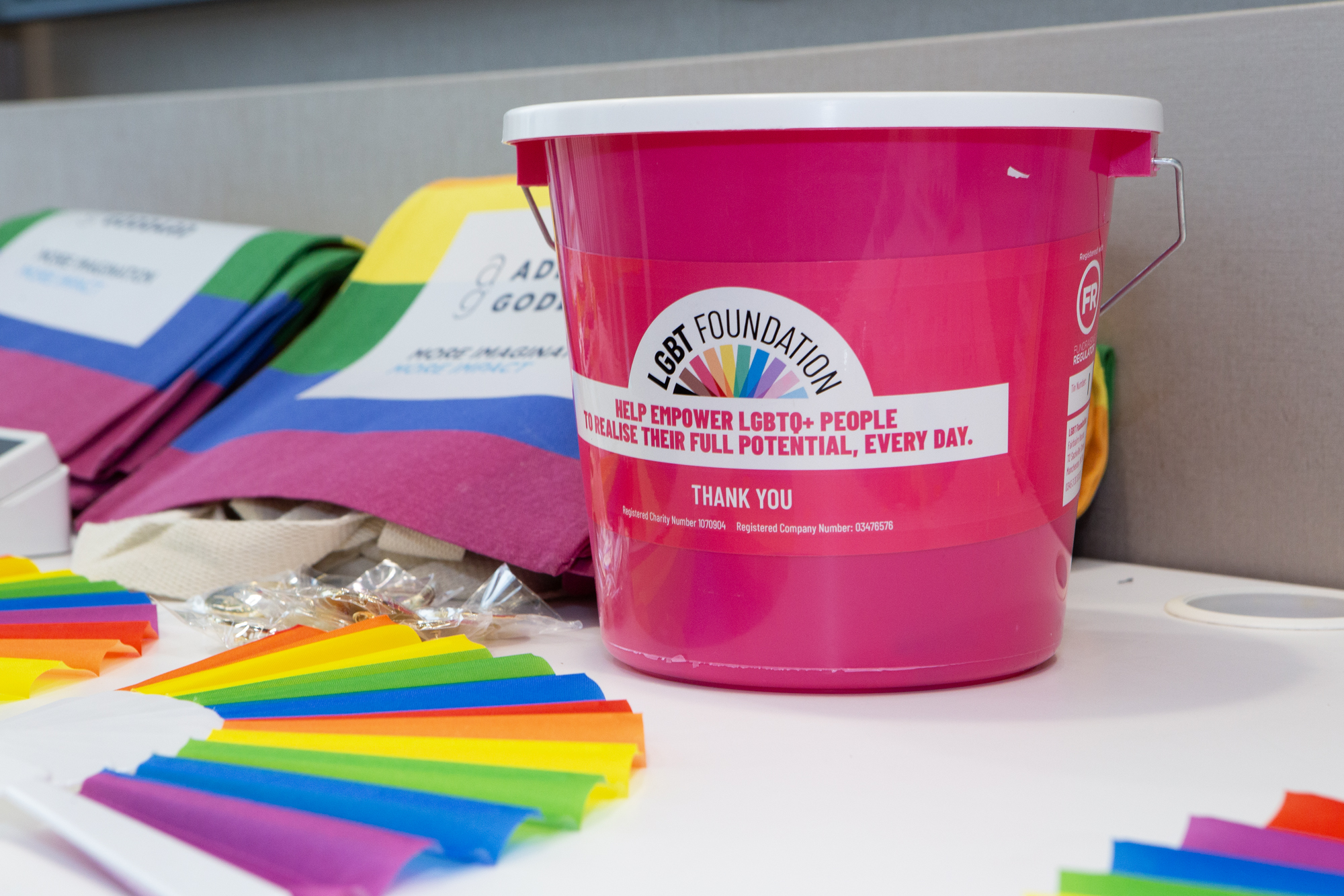 A large pink LGBT Foundation donation bucket is the focus of this image. In the background is more rainbow-themed merchandise on a table.