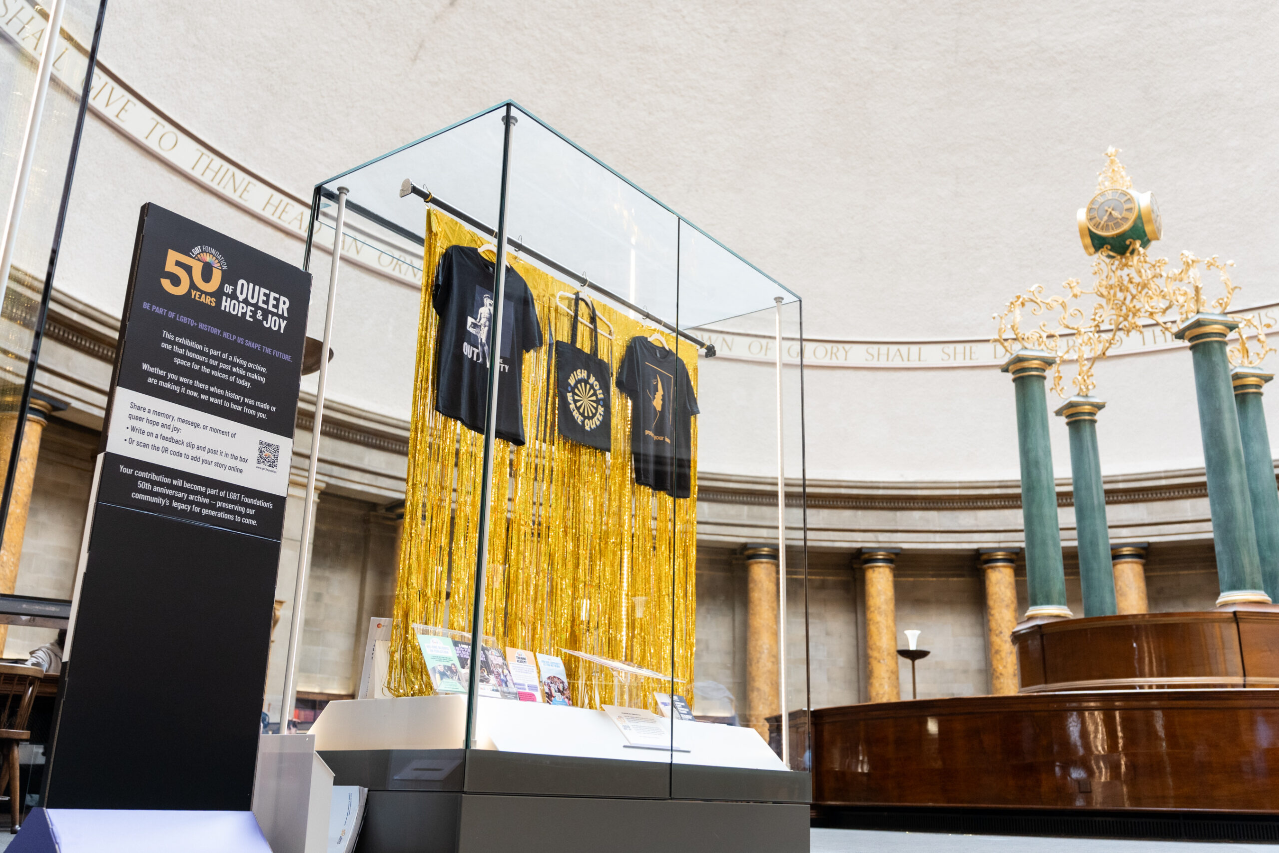 A selection of LGBT Foundation's 50th Anniversary designed merchandise hung inside a glass display cabinet with a shiny gold backdrop. To the left of the cabinet is an information board about LGBT Foundation's 50th Anniversary.