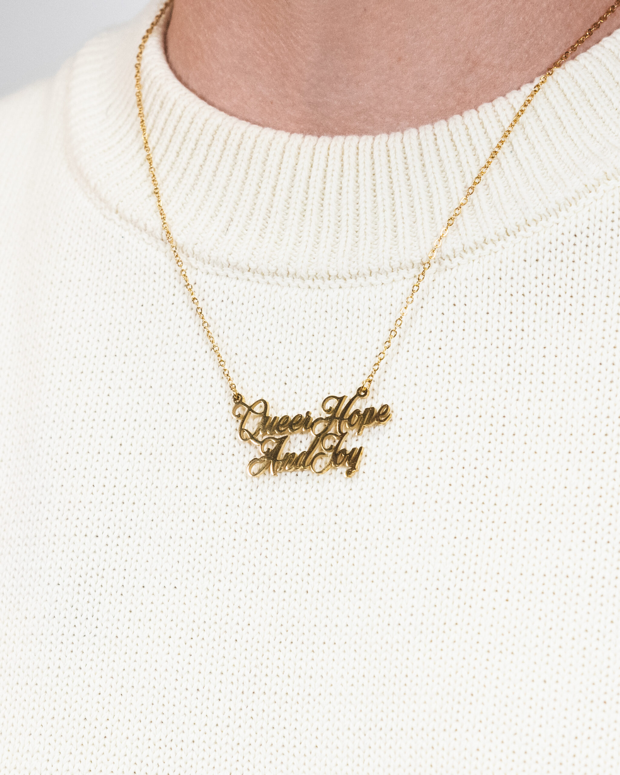 Close-up photo of a gold "Queer Hope & Joy" necklace around a person's neck, against a cream jumper.
