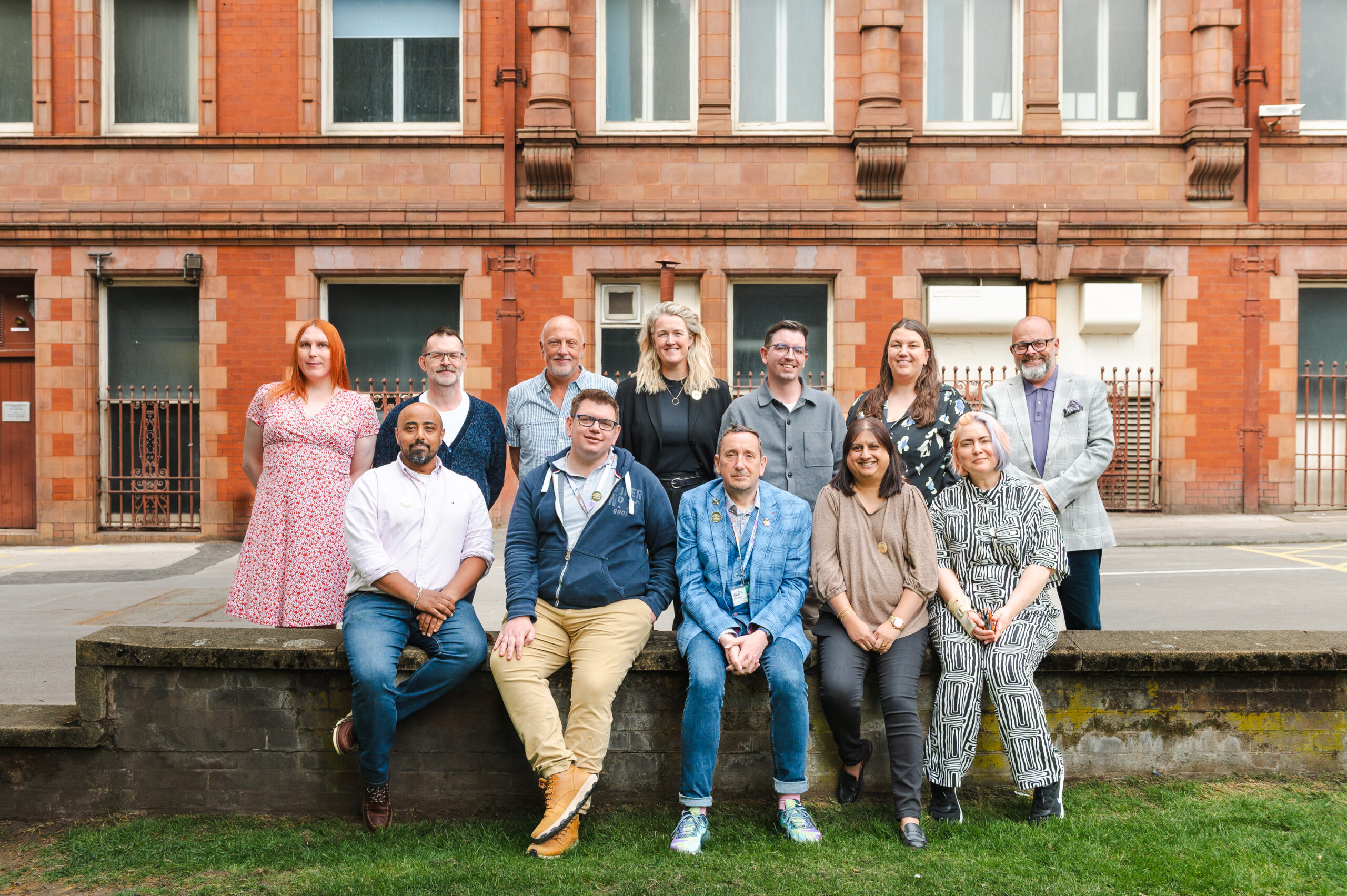A photograph of LGBT Foundation's trustees in two rows, one standing and one seated on a wall in front of a building in Manchester.