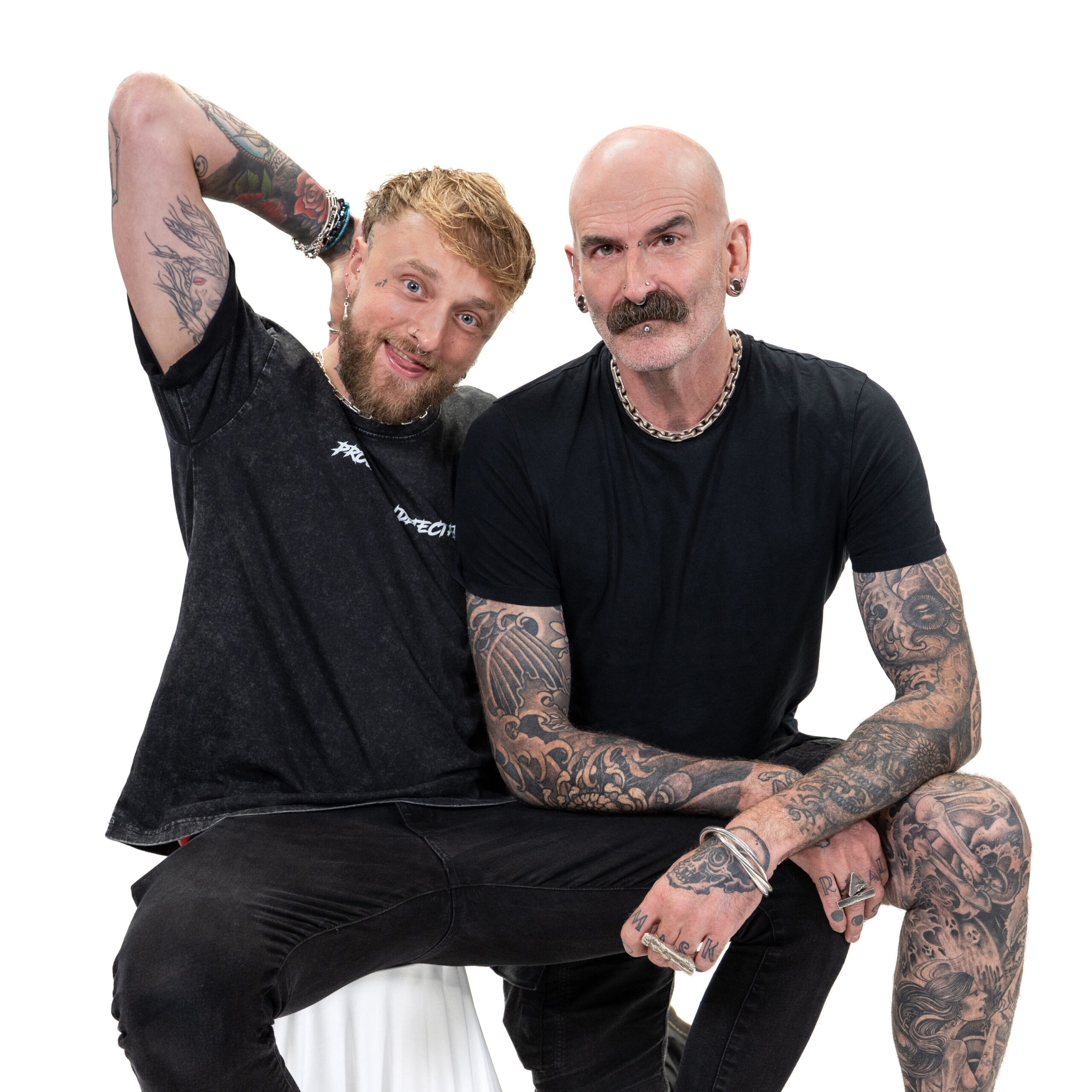 A pair of LGBTQ+ men with tattoos dressed in black, smiling and looking at the camera against a white backdrop for LGBT Foundation's Man Enough campaign