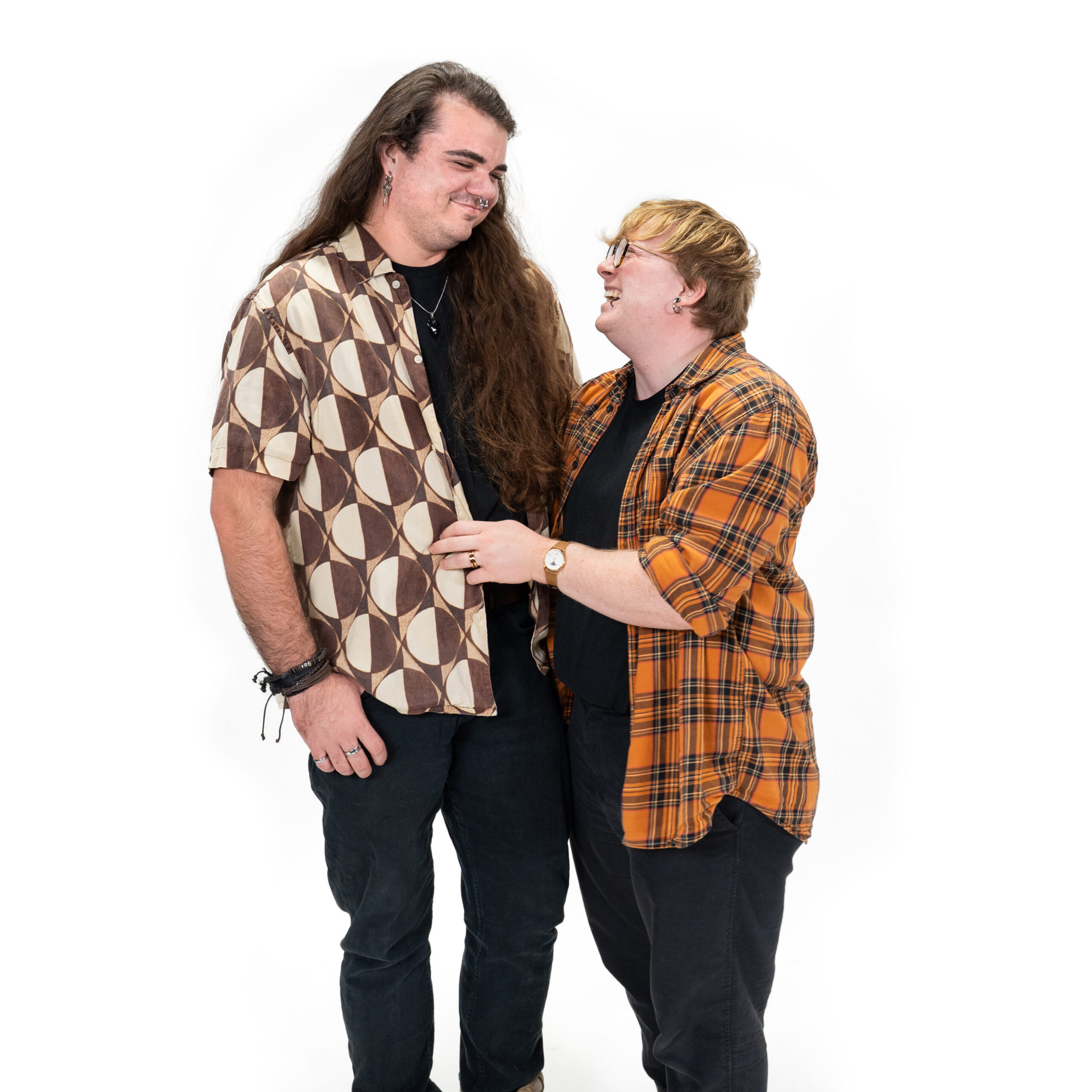A trans+ couple wearing complementary checkered shirts smiling and looking at each other against a white background.